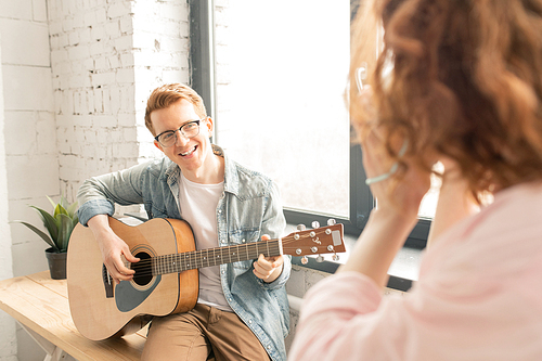Happy young singing man playing the guitar while looking in smartphone camera held by girl recording home party