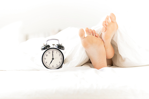 Alarm clock on bed next to bare female feet and soles stuck out of white blanket or sheet during sleep