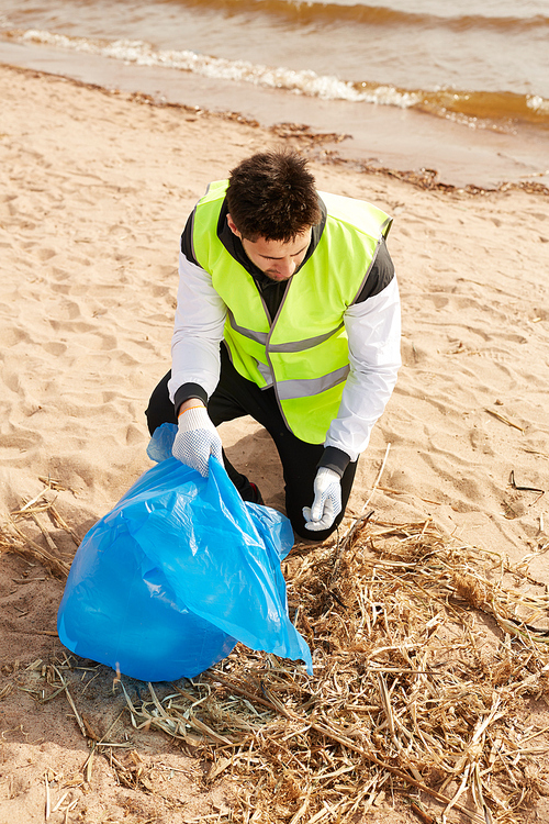 Young contemporary volunteer in uniform picking up wastes while working on sandy beach