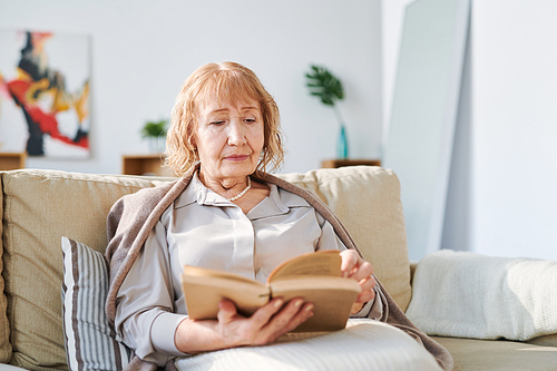 Senior female in casualwear sitting on comfortable couch by cushion and reading book of stories or novel in living-room