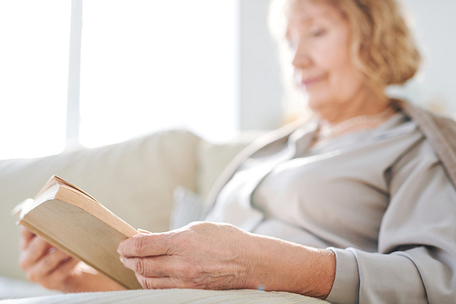 Hands of senior female pensioner holding open book while reading it while relaxing on sofa at leisure