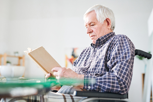aged handicapped man in checkered shirt sitting in . by table and reading novel at leisure in retirement home