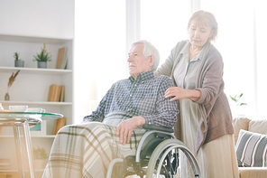 Mature female caregiver in casualwear taking care of aged disable man with grey hair in wheelchair