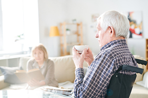Senior man in wheelchair having tea in retirement home on background of mature woman reading to him