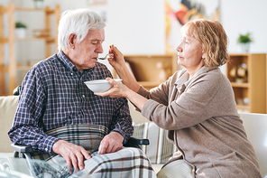 Aged casual woman holding bowl with soup and spoon by her disable husband mouth while helping him to eat dinner