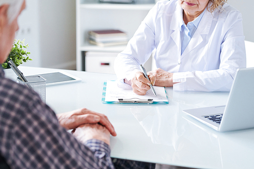 Mature doctor making prescriptions on paper while sitting by desk in front of aged handicapped patient in clinics