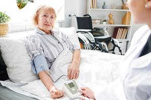 Sick aged female patient of therapy department sitting in bed and listening to doctor measuring her blood pressure