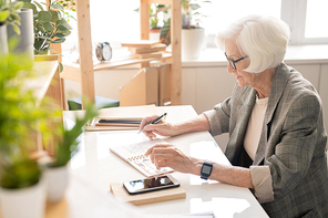 Senior white-haired businesswoman in formalwear sitting by desk in office and reading notes in notebook while planning work