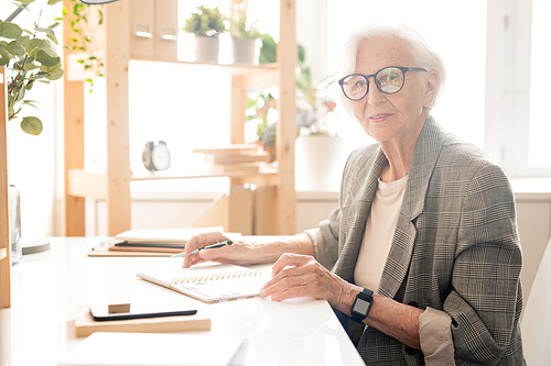 Successful aged female in eyeglasses and formalwear looking at you while sitting by desk and organizing work in office