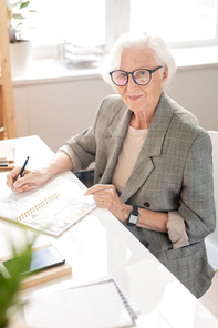 Successful aged office worker with white hair sitting by desk while planning working day in the morning