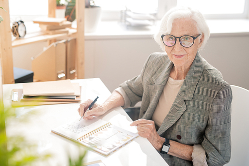 Elderly businesswoman with white hair writing down plan of working day in notebook by desk in front of camera