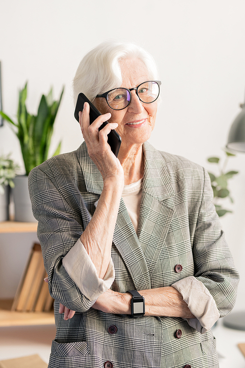 Aged happy white-haired female in eyeglasses and formalwear standing in front of camera while talking on smartphone