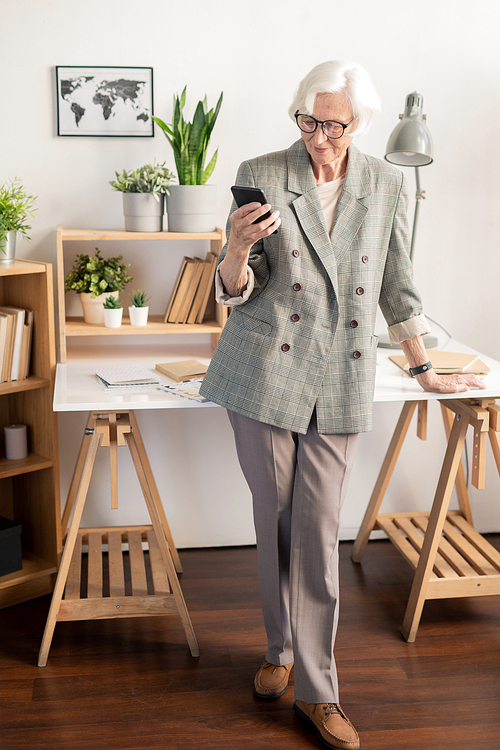 Elegant retired white-haired woman in grey suit texting in smartphone while standing by desk with working supplies