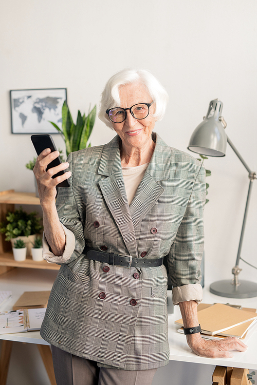 Successful senior businesswoman with mobile gadget looking at you while leaning by desk with working supplies