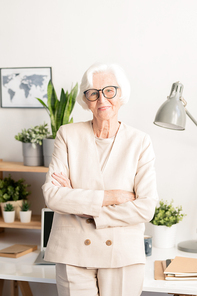 Happy aged elegant woman in white suit and eyeglasses crossing her arms on chest while standing in front of camera