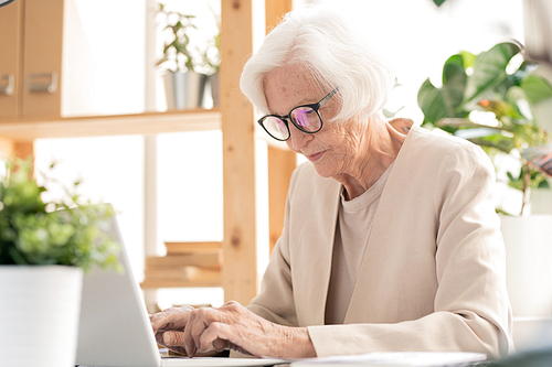 Senior busy elegant female in white suit and eyeglasses typing on laptop keypad while browsing in the net by desk