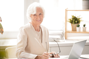 Aged smiling businesswoman in white elegant suit looking at you while sitting by desk in front of laptop in office