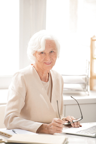 Aged successful female business leader in white elegant suit sitting by workplace in front of laptop during work