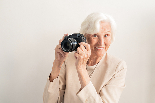 Mature white-haired smiling female photographer with photocamera by face standing on light background