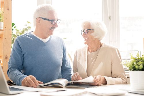 Happy senior couple in smart casualwear looking at each other while sitting by desk and looking through magazine