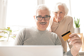 Contemporary senior man with plastic card sitting in front of laptop while shopping online and paying for orders