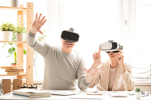 Two contemporary seniors in vr headsets sitting by desk while touching virtual display during presentation