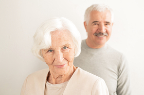 Smiling senior female with white hair looking at you while standing in front of camera with her husband on background