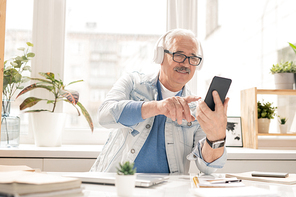 Creative aged man in casualwear and headphones looking through playlist while sitting by workplace in office