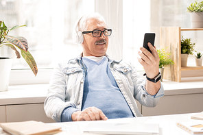 Contemporary mature mobile man in casualwear and headphones watching video in smartphone while sitting in office