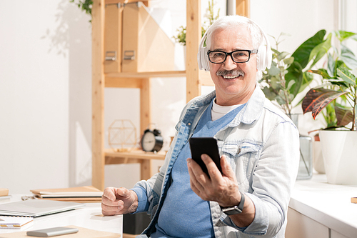 Successful aged man in headphones and casualwear using smartphone while sitting by desk in front of camera