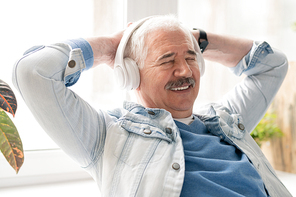Mature smiling relaxed man enjoying music in headphones while keeping his hands on back of head in the office