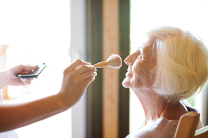 Contemporary beautician with powder brush working over makeup of senior lady before photo shooting