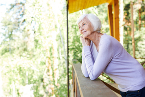Happy serne senior female in casualwear enjoying rest in the country house while standing by wooden banisters