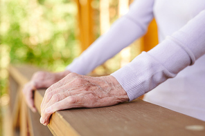 Hands of senior woman in white long-sleeved pullover holding by wooden banisters of her country house during relax