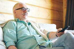 Restful senior man in eyeglasses and casualwear using remote control while watching tv in his country house