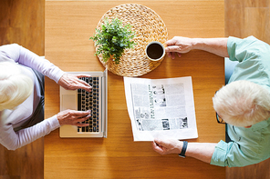 Overview of senior husband with newspaper and wife with laptop sitting by wooden table in front of one another