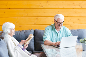 Restful senior spouses sitting on sofa in living-room, husband networking in front of laptop and wife reading book