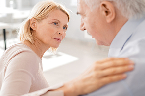 Compassionate aged blonde female looking at suffering senior man while keeping her hand on his shoulder
