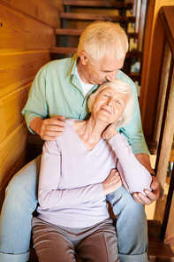 Senior man kissing his wife head and embracing her while both sitting on wooden staircase at home