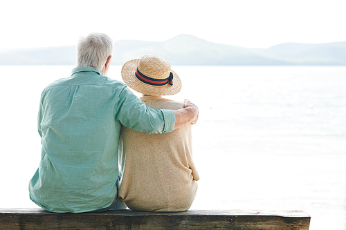 Back view of serene senior couple in casualwear looking at lake while relaxing by waterside on summer day
