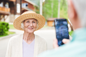 Happy senior female in elegant straw hat posing for camera while standing in front of her husband taking photograph on smartphone
