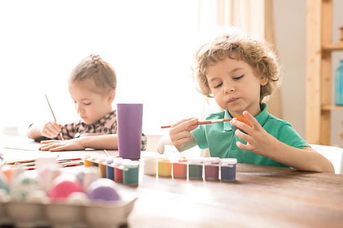 Adorable boy with curly hair painting eggs with gouache by table on background of his sister