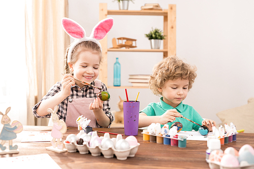 Happy little girl with soft pink rabbit ears headband and her brother painting eggs for Easter festival