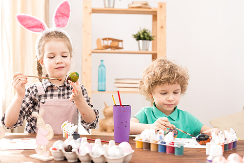Adorable little girl with Easter bunny headband painting eggs for Easter with her younger brother near by