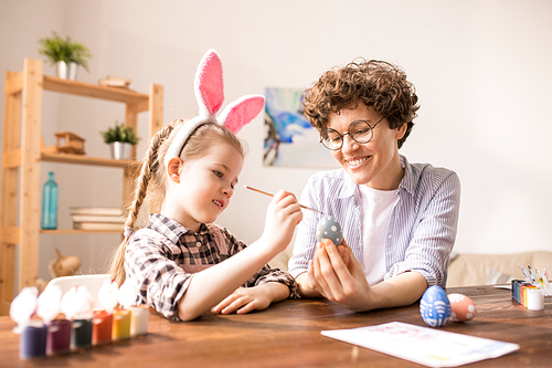 Young mother helping little girl with painting Easter eggs while both sitting by table at home