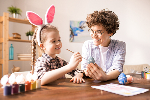 Smiling girl applying white paint on grey Easter egg held by her mother while both sitting by table at leisure