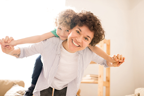 Young cheerful woman in casualwear giving piggyback to her little son while having fun at home