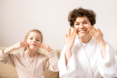 Pretty little daughter and her young mother taking care of skin - applying cream on their faces and massaging gently