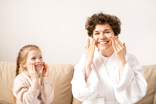Young woman with gorgeous smile applying cream on face while little daughter repeating after her