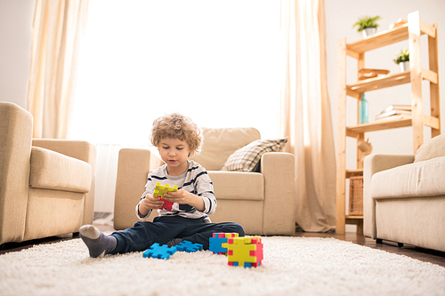 Little boy sitting on the floor of living-room and making construction of colorful parts at leisure